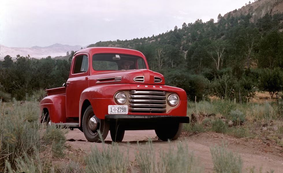 A classic red 1948 Ford F-1 pickup truck parked on a dirt path surrounded by greenery.