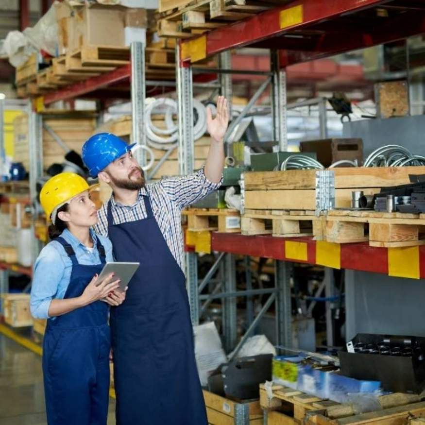 Two workers in hard hats and aprons inspect shelves in an aftermarket auto parts store warehouse.