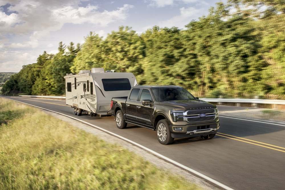 A Ford F-150 truck towing a large camper trailer on a scenic road with trees and greenery in the background.