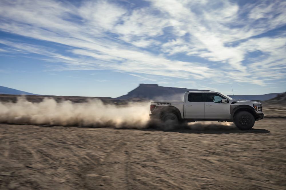 A white Ford F-150 kicks up dust as it speeds through a desert landscape under a blue sky.