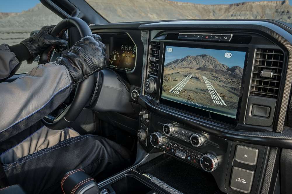 A driver in gloves operates a Ford F-150, with the dashboard screen displaying a rugged landscape.