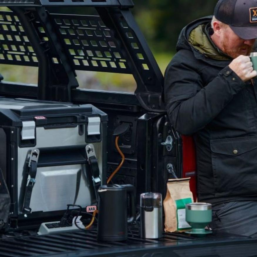 A man enjoys coffee by the spacious interior of a Toyota Tacoma's tailgate, equipped with camping gear for an outdoor adventure.