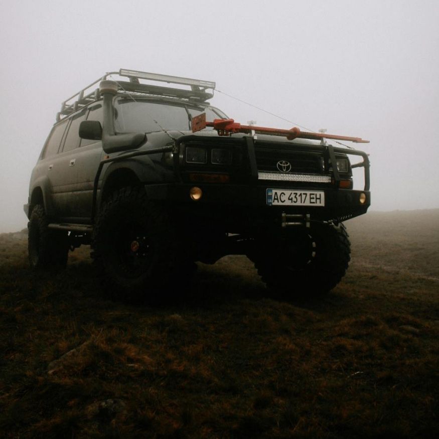 Off-road black Toyota Land Cruiser on rugged terrain in dense fog, equipped with roof rack and front winch.