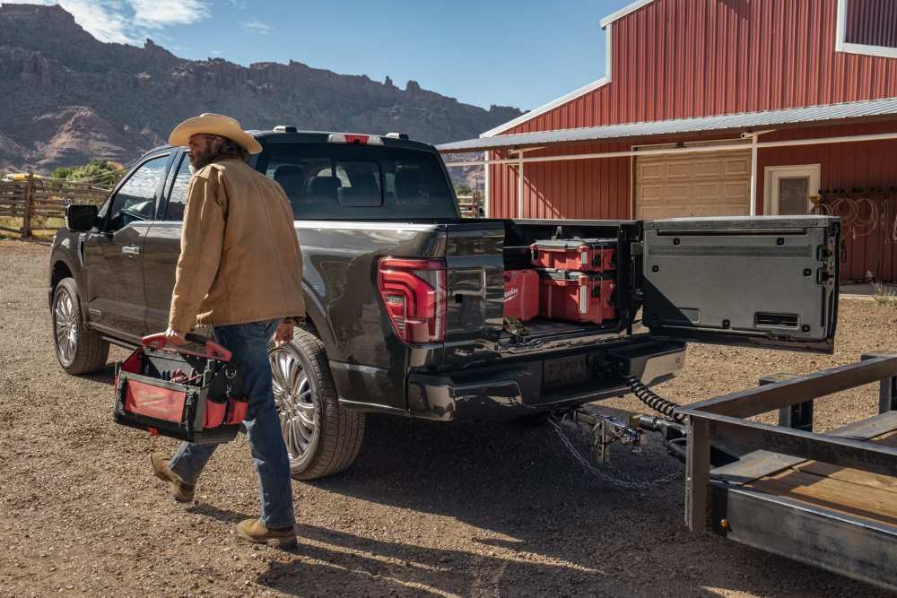 Man in cowboy hat carrying a maintenance toolbox near a Ford F-150 truck with an open tailgate, parked by a red barn in a rural setting.