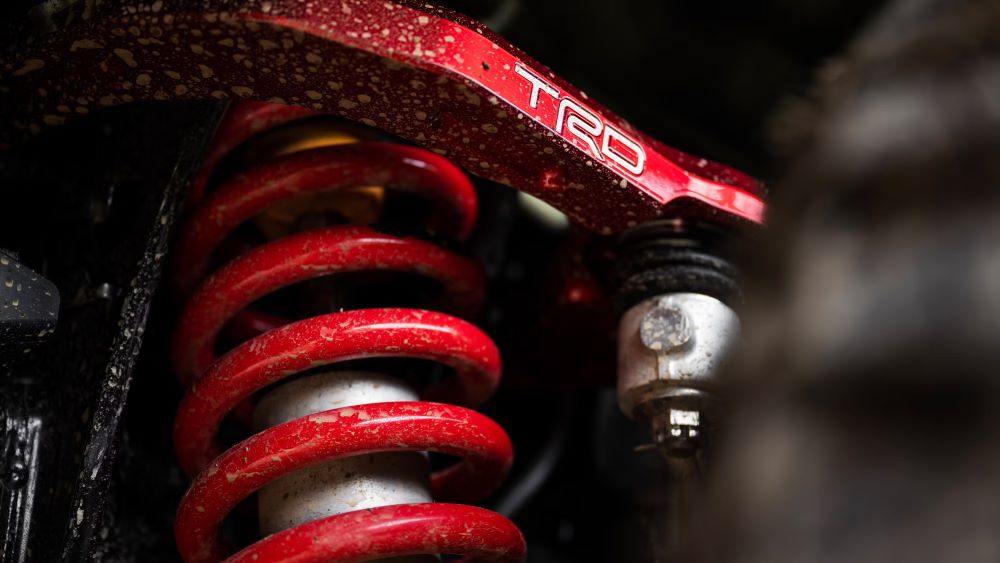 Close-up of a Toyota Tacoma TRD suspension upgrade with red springs and a TRD-branded component, showcasing off-road performance enhancements.