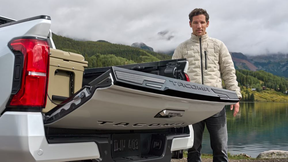 Man beside a silver 2024 Toyota Tacoma with an open tailgate near a lake and mountains.