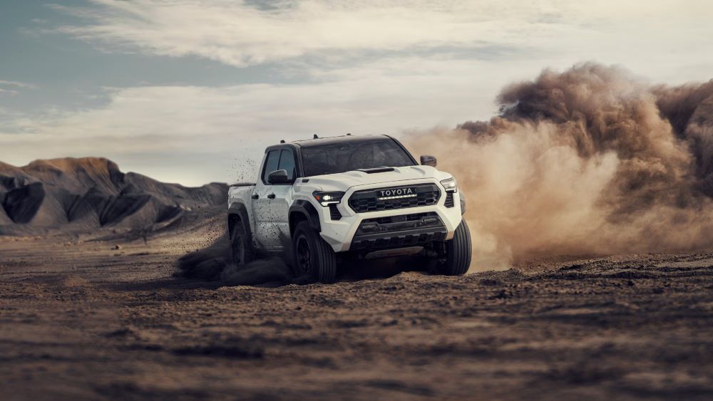 A white 2024 Toyota Tacoma driving through dirt with dust clouds trailing behind, showcasing its rugged design and off-road capability.