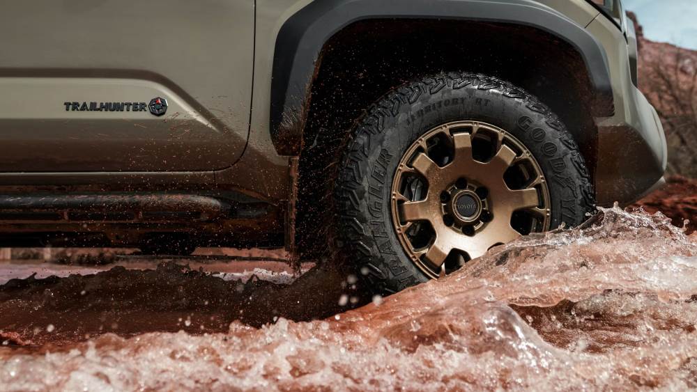 Close-up of a Toyota Tacoma Trailhunter driving through muddy water, showcasing the Trailhunter badge and Goodyear tires.