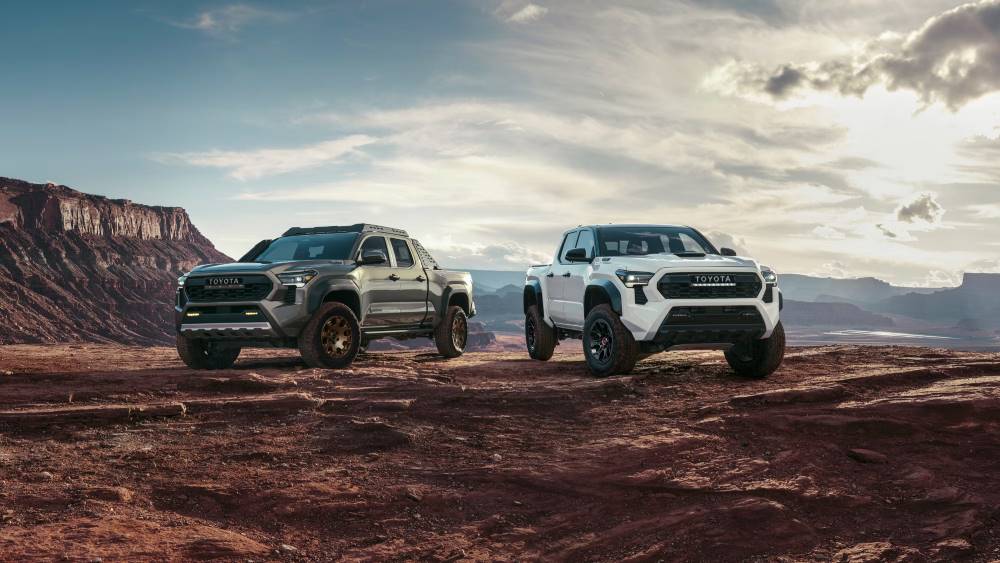 The image shows two rugged Toyota Tacoma trucks in a desert setting, with mountains in the background, highlighting their off-road capabilities.