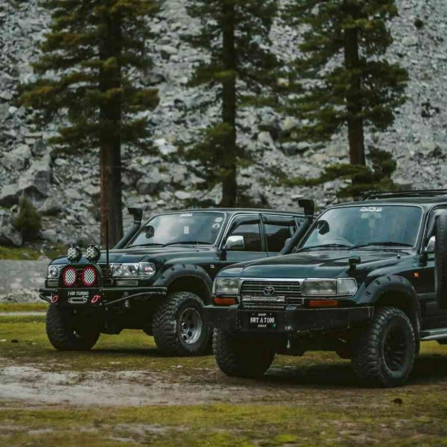 Two upgraded black Toyota Land Cruisers parked on grass, with pine trees and rocky hills behind.